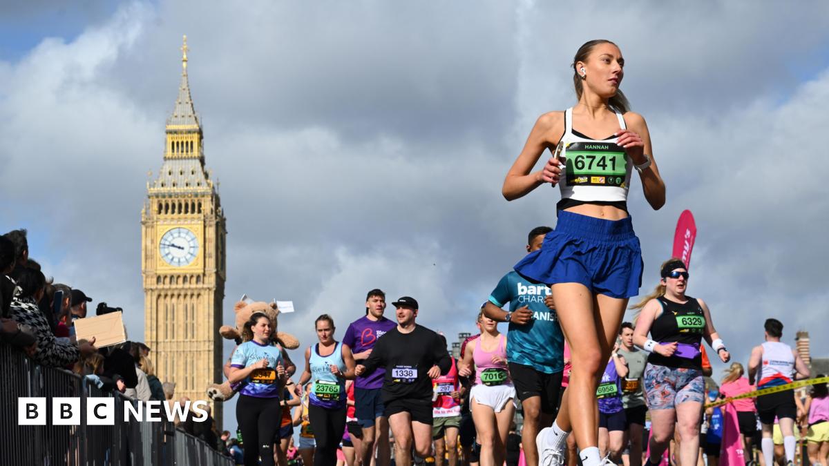 A large group of people participate in the London Landmarks Half Marathon, running on a city street with the Elizabeth Tower visible in the background under a cloudy sky.