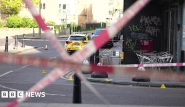 Red police tape crosses the road. Silver chairs are on the pavement opposite, inside the police cordon with a police car with fluorescent yellow marking parked beyond it.
