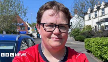 A person wearing a red England football shirt with short hair and glasses is standing on a residential street
