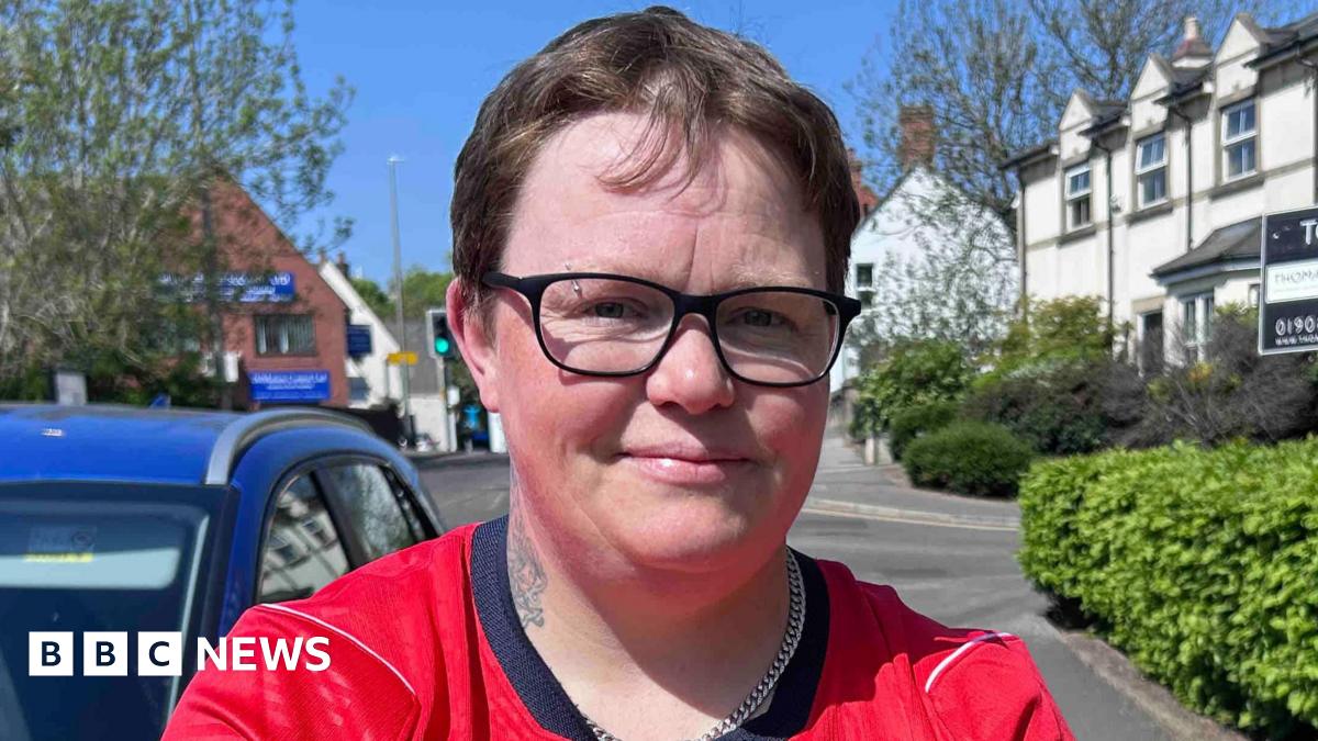 A person wearing a red England football shirt with short hair and glasses is standing on a residential street