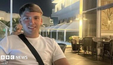 Nathan sitting in a pub with a drink. He is wearing a blue tracksuit top and has his arms crossed. He has a short beard and dark brown hair.