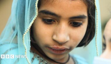 A woman and a girl sit on a bench made of metal and woven fabric. The woman wears a dark veil that covers her face and head, with her eyes visible, while the girl is wearing light blue clothes and a scarf over the top of her head