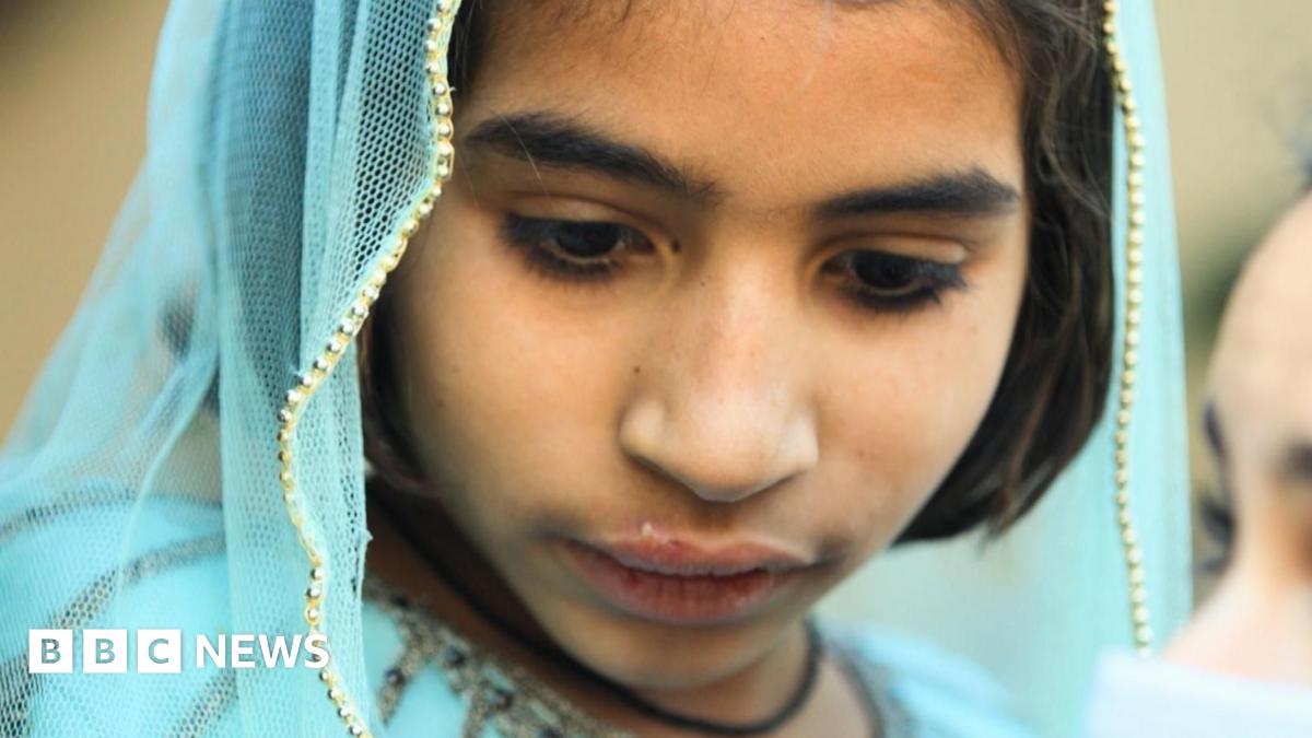 A woman and a girl sit on a bench made of metal and woven fabric. The woman wears a dark veil that covers her face and head, with her eyes visible, while the girl is wearing light blue clothes and a scarf over the top of her head