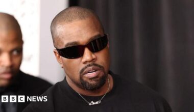 Headshot of West who wears a silver chain and sunglasses. There is a man behind him who is blurred