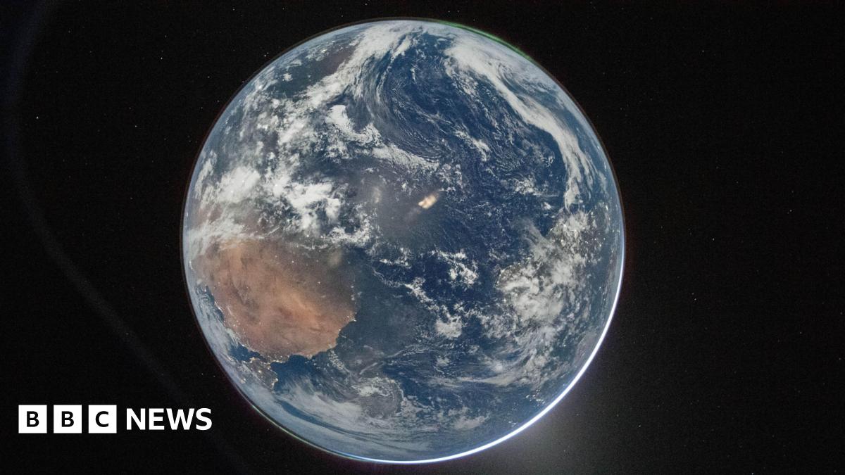 A picture of the Earth from space, which centres the planet against the dark background of space. It's a round blue planet. Clouds can be seen and a thin green aurora at the top.