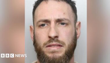 A mugshot of a man with blue eyes, a moustache and beard