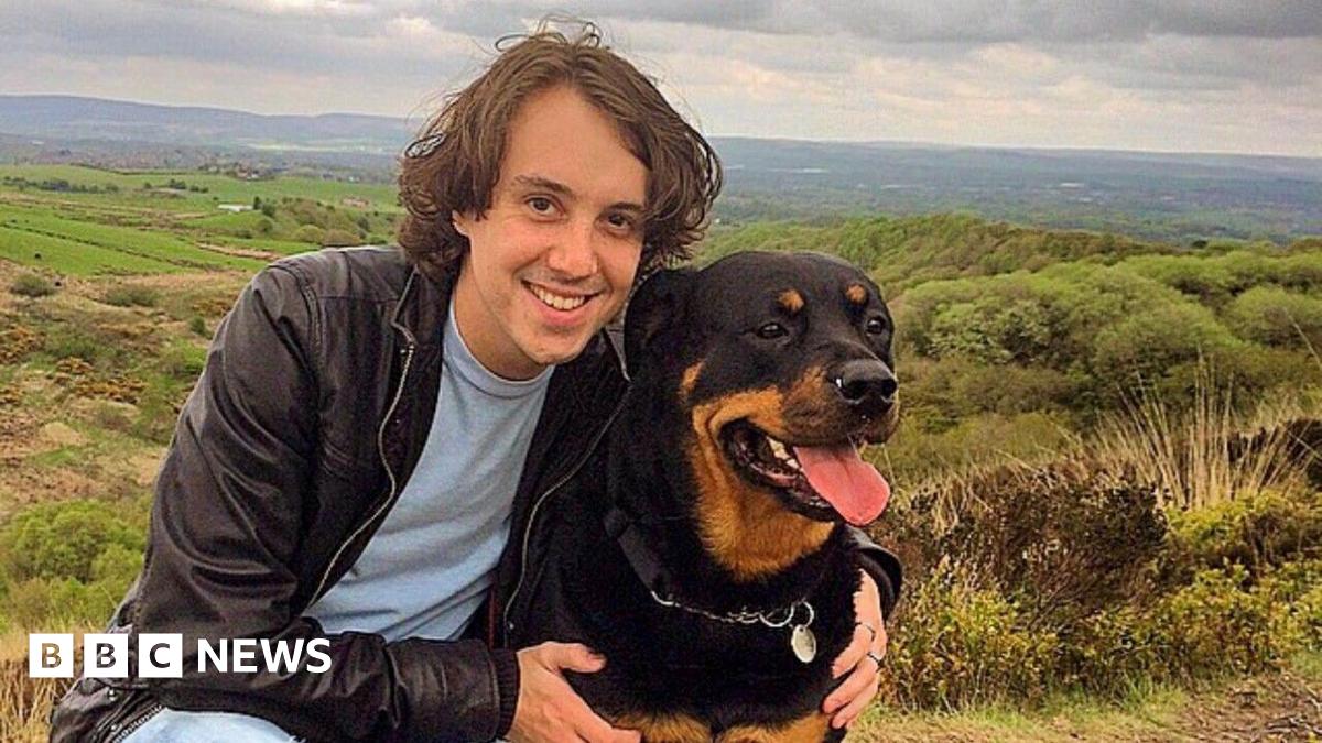 Oliver Robinson with medium length brown hair, wearing a dark jacket and light blue tshirt smiles at the camera with his arms around a black and tan rottweiler dog with countryside and fields in the background