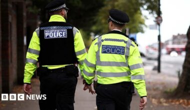 Two Metropolitan Police officers patrol a street in hi-vis jackets. Their backs are turned towards the camera.