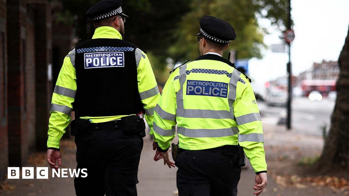 Two Metropolitan Police officers patrol a street in hi-vis jackets. Their backs are turned towards the camera.