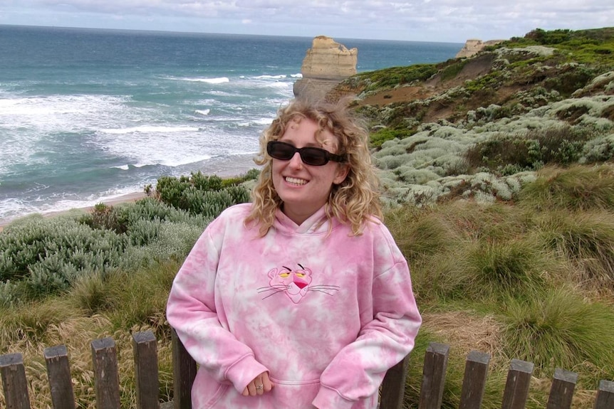 Woman in pink hoodie and sunglasses smiles at camera. she has blonde curly hair. Ocean is in the background