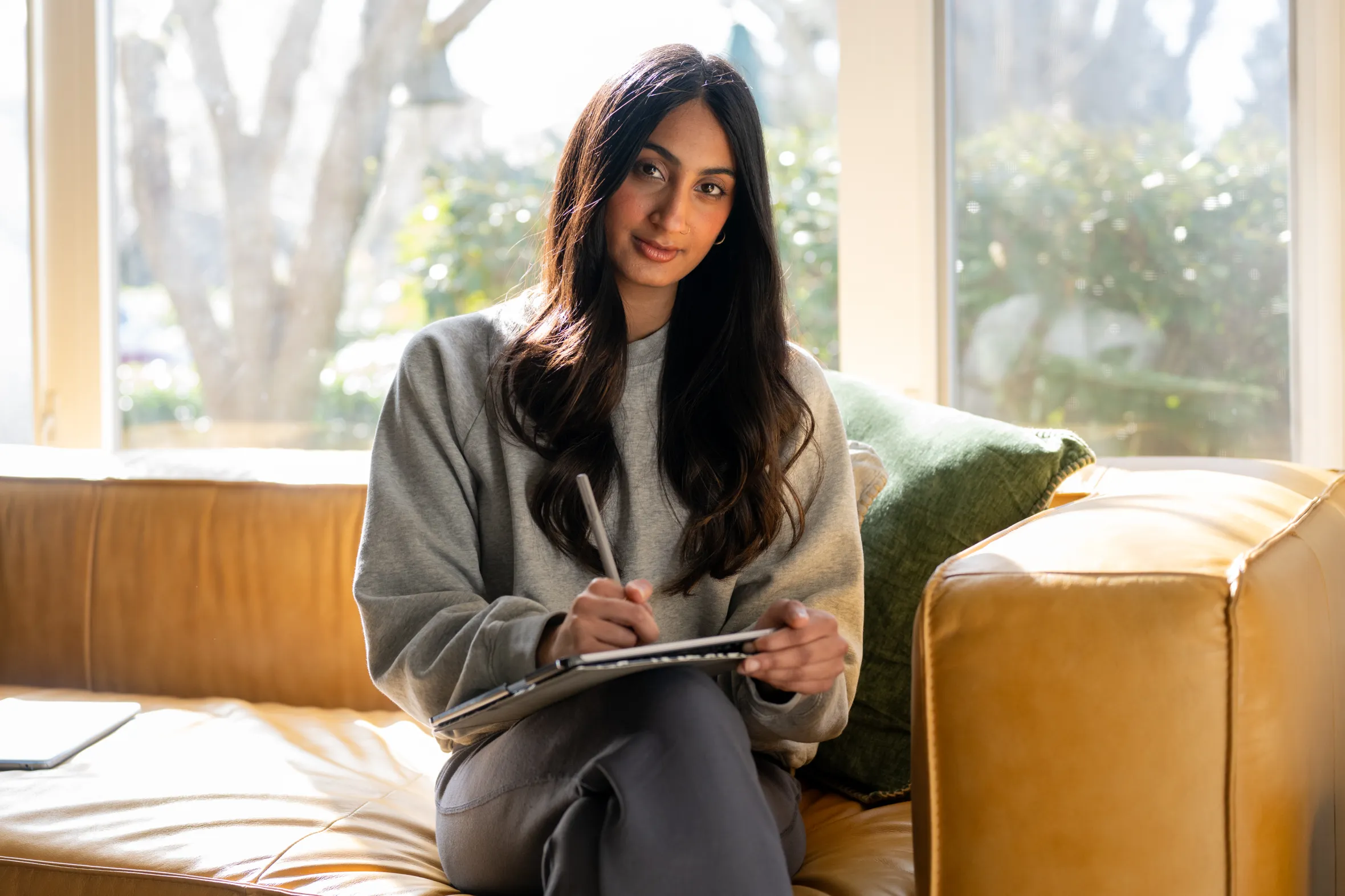 Thea Bhoopalan seated on a brown couch, holding a pen and notebook.