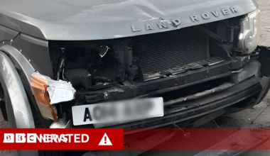 A close-up of the damaged front of a grey Land Rover, with extensive damage to the bumper and lights of the vehicle. A white, partly-blurred number plate is positioned on the left of the bumper.