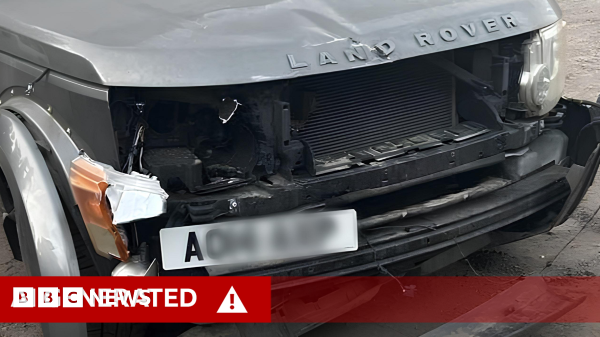 A close-up of the damaged front of a grey Land Rover, with extensive damage to the bumper and lights of the vehicle. A white, partly-blurred number plate is positioned on the left of the bumper.