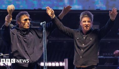 Noel and Liam Gallagher, both wearing black tops and jeans, raise their hands in the air on stage during their reunion tour on stage. Liam is holding maracas in his right hand.