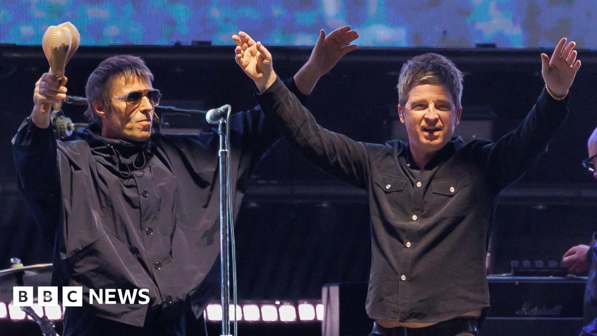 Noel and Liam Gallagher, both wearing black tops and jeans, raise their hands in the air on stage during their reunion tour on stage. Liam is holding maracas in his right hand.