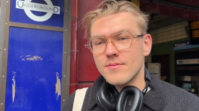A man with glasses and blond hair stands in front of an Underground station entrance.