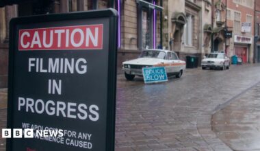 Two vintage Rover police cars, white with a red stripe across the side and blue light on top, parked up outside a row of grand-looking early 20th Century buildings, including the former Bob Carver's chip shop. A sign reads "police slow". In the foreground, another sign reads "caution, filming in progress, apologies for any inconvenience caused".