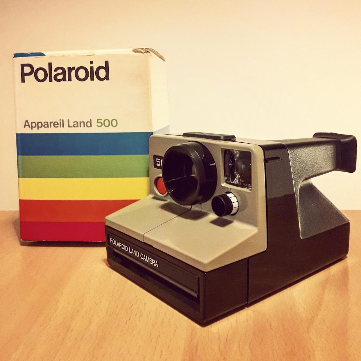 A vintage Polaroid Land Camera sits on a wooden surface next to its original box, which features the Polaroid logo and colorful stripes.