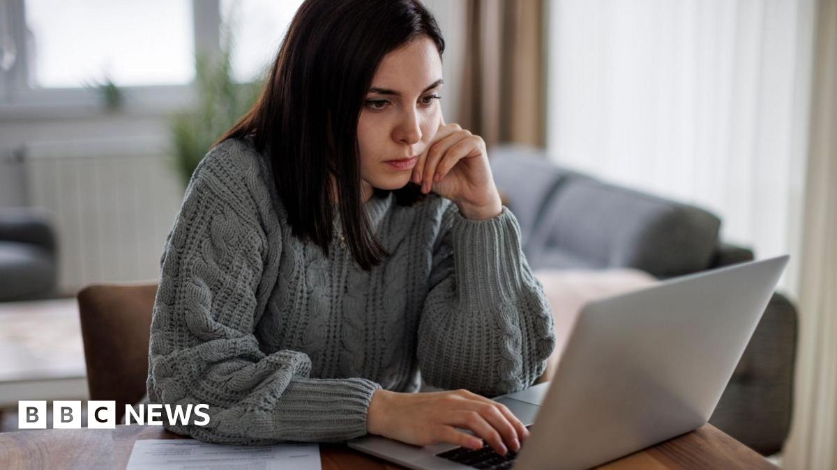 Woman with long dark hair and wearing a grey jumper sits at a desk looking at a laptop computer