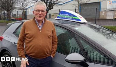 A man with white hair and glasses standing smiling in front of a learners car parked on a street by an industrial estate. He is wearing a brown top and navy bottoms.