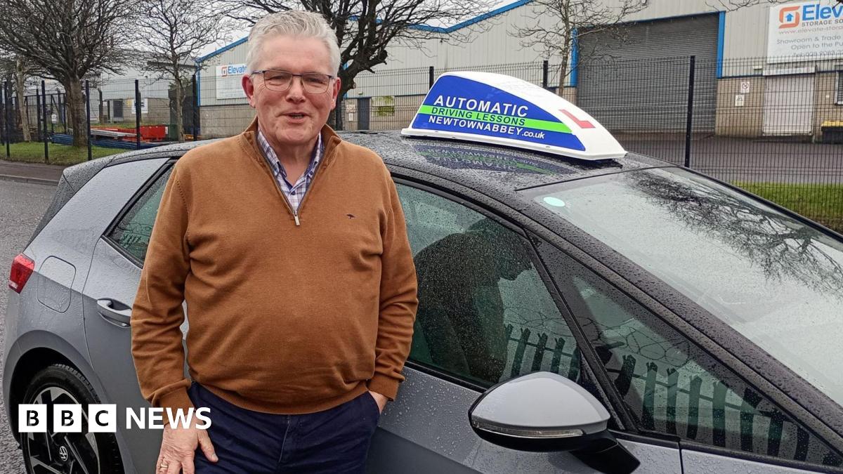 A man with white hair and glasses standing smiling in front of a learners car parked on a street by an industrial estate. He is wearing a brown top and navy bottoms.