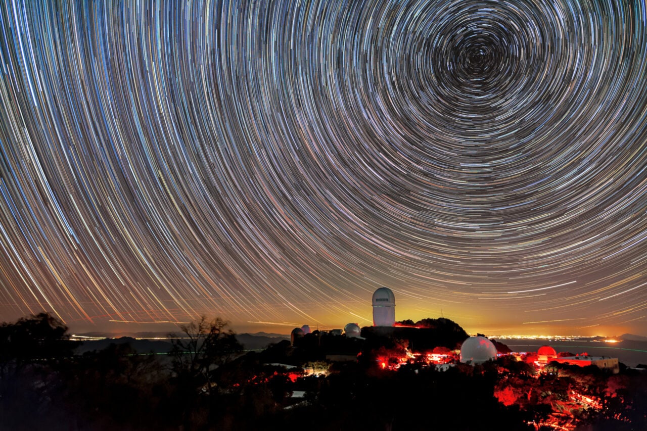 Circles of light on the night sky. A telescope dome atop a mountain is below the center of the circle.