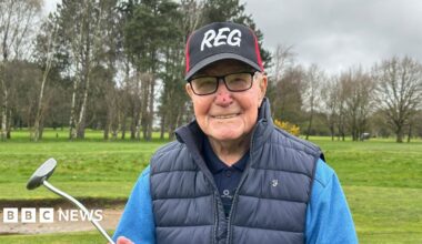 A man with black rimmed glasses is pictured smiling whilst holding a golf putter with a black and red cap featuring his name, Reg, in white capital letters.  A golf course is in the background. He's also sporting a navy gilet over a light blue jumper and navy blue polo shirt.