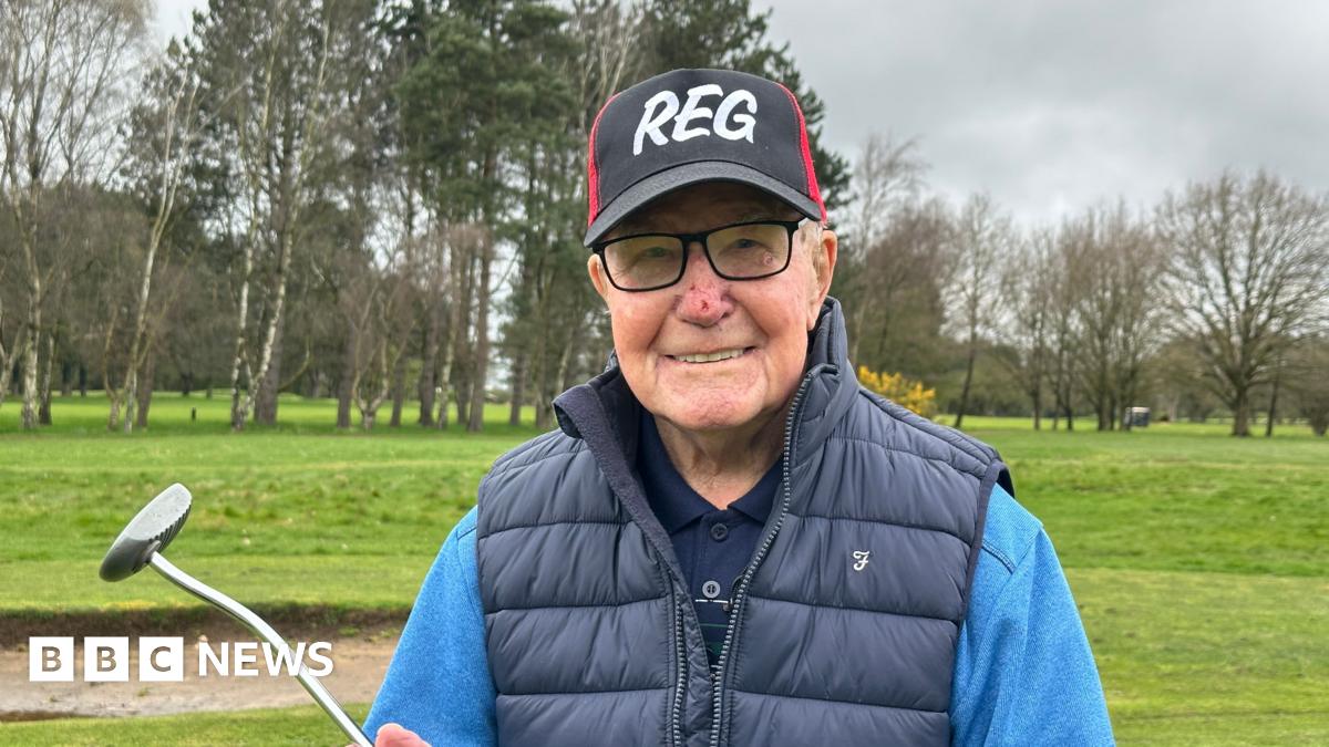 A man with black rimmed glasses is pictured smiling whilst holding a golf putter with a black and red cap featuring his name, Reg, in white capital letters.  A golf course is in the background. He's also sporting a navy gilet over a light blue jumper and navy blue polo shirt.
