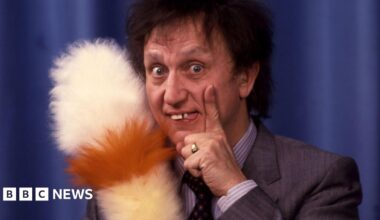 Ken stands with his trademark feather duster in hand in front of a draped curtain. His hand rests on his face. He's wearing a suit.
