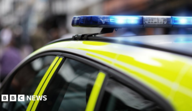 The top of a yellow police vehicle with the blue lights above the vehicle being on. The background is blurred.