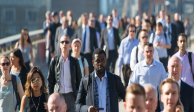 men and women mostly smartly dressed in shirts and jackets walking towards the camera seemingly in London over a bridge in the sunshine