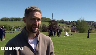 Woakes with brown hair and a brown top is on the left of the photo. A field and other people are in the background, while trees and rooftops are in the distance.