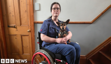 A woman with tied-back brown hair is wearing a dark blue surgical gown and sitting in a red wheelchair. She has a small terrier-type dog on her lap. They are in a corridor with wooden flooring and stairs.