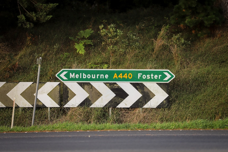 Green and white road sign pointing to Melbourne in one direction and Foster in the other, with green hil behind