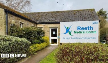 A GP practice which is a single storey building with a glass door and hedges around the entrance. A sign reads Reeth Medical Centre.