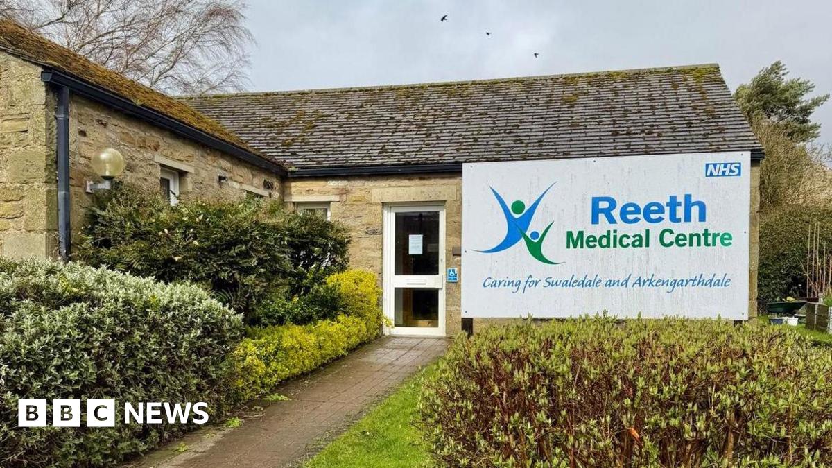 A GP practice which is a single storey building with a glass door and hedges around the entrance. A sign reads Reeth Medical Centre.