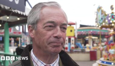 Nigel Farage, who is wearing a green wax jacket and shirt, smiles while stood outside at an amusement park. Rides, including a carousel, can be seen in the background.