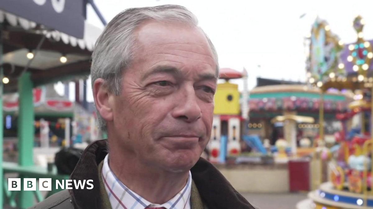 Nigel Farage, who is wearing a green wax jacket and shirt, smiles while stood outside at an amusement park. Rides, including a carousel, can be seen in the background.
