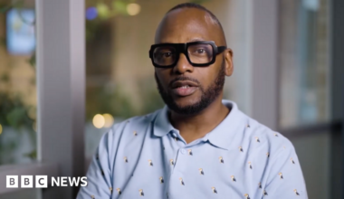 Christopher Joell-Deshields looks at the camera, his lips pursed as though he is about to speak. He is wearing a blue polo shirt covered in embroided puffins. The background is out of focus