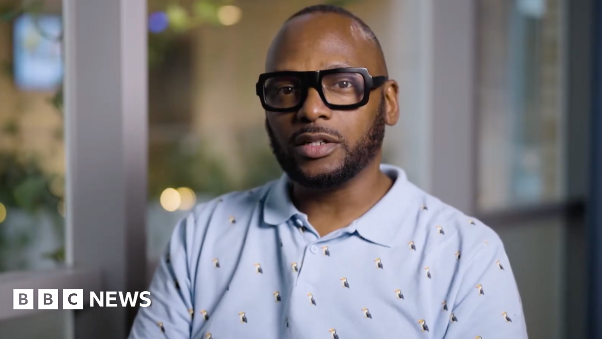 Christopher Joell-Deshields looks at the camera, his lips pursed as though he is about to speak. He is wearing a blue polo shirt covered in embroided puffins. The background is out of focus