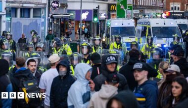 A large group of people standing near riot police, who are armed with shields and helmets. There are several police vans parked nearby some shops along a high street.