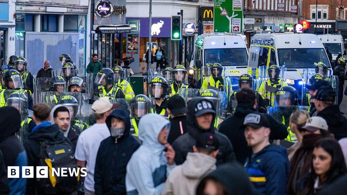 A large group of people standing near riot police, who are armed with shields and helmets. There are several police vans parked nearby some shops along a high street.