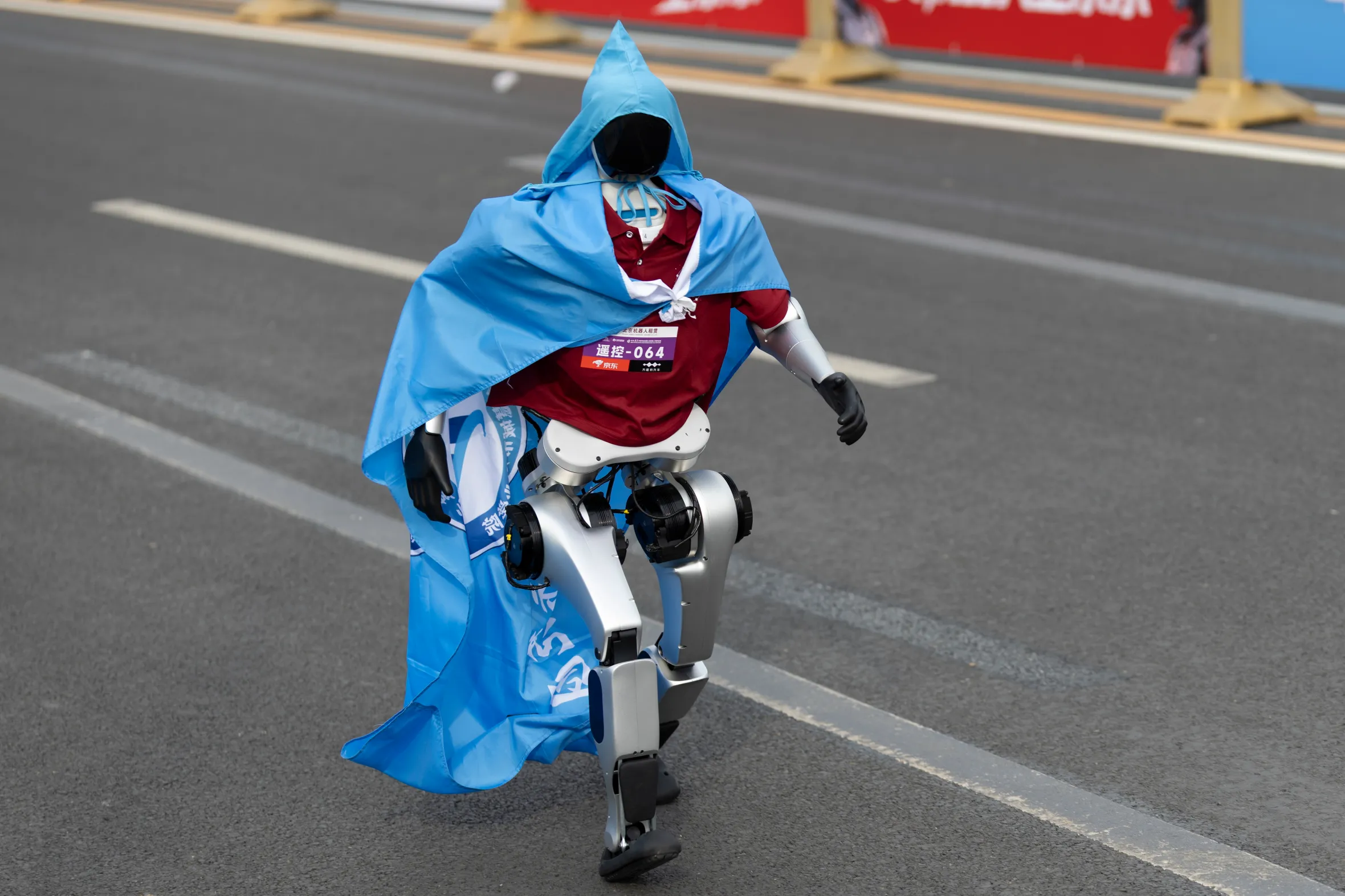 A humanoid robot dressed in a maroon shirt and blue cape runs on a paved road.