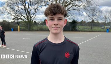 Oscar is stood in the school playground in a school branded black top with red piping and a red logo. He has mousy coloured, curly hair, and his hands behind his back.