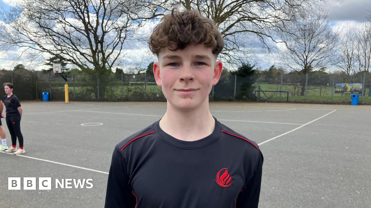 Oscar is stood in the school playground in a school branded black top with red piping and a red logo. He has mousy coloured, curly hair, and his hands behind his back.