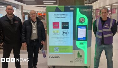 Three people stand inside a busy shopping centre beside a bright EcoVend reverse‑vending machine, which collects empty plastic bottles and cans. The group appear to be staff or project partners, wearing uniforms and lanyards as they pose for the photo. Behind them, high‑street shops and food outlets form the backdrop, emphasising the machine’s prominent position in a public retail space. The EcoVend unit displays messaging about turning recyclables into in‑store rewards, along with logos of participating local businesses.