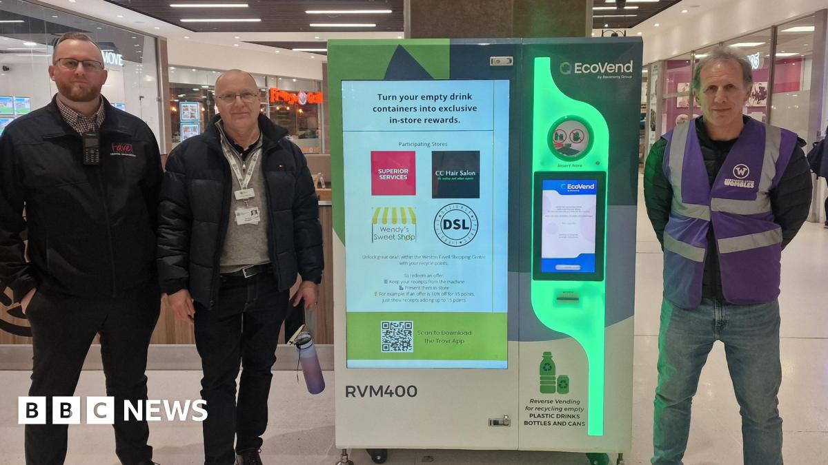 Three people stand inside a busy shopping centre beside a bright EcoVend reverse‑vending machine, which collects empty plastic bottles and cans. The group appear to be staff or project partners, wearing uniforms and lanyards as they pose for the photo. Behind them, high‑street shops and food outlets form the backdrop, emphasising the machine’s prominent position in a public retail space. The EcoVend unit displays messaging about turning recyclables into in‑store rewards, along with logos of participating local businesses.