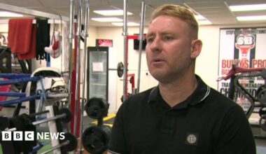 Andrew Bulcroft pictured in a gym with weight equipment behind him. He has short blonde hair and is wearing a black polo shirt.