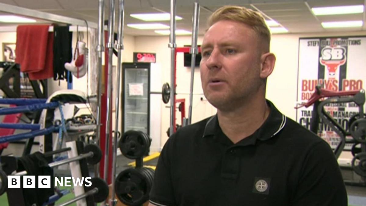 Andrew Bulcroft pictured in a gym with weight equipment behind him. He has short blonde hair and is wearing a black polo shirt.