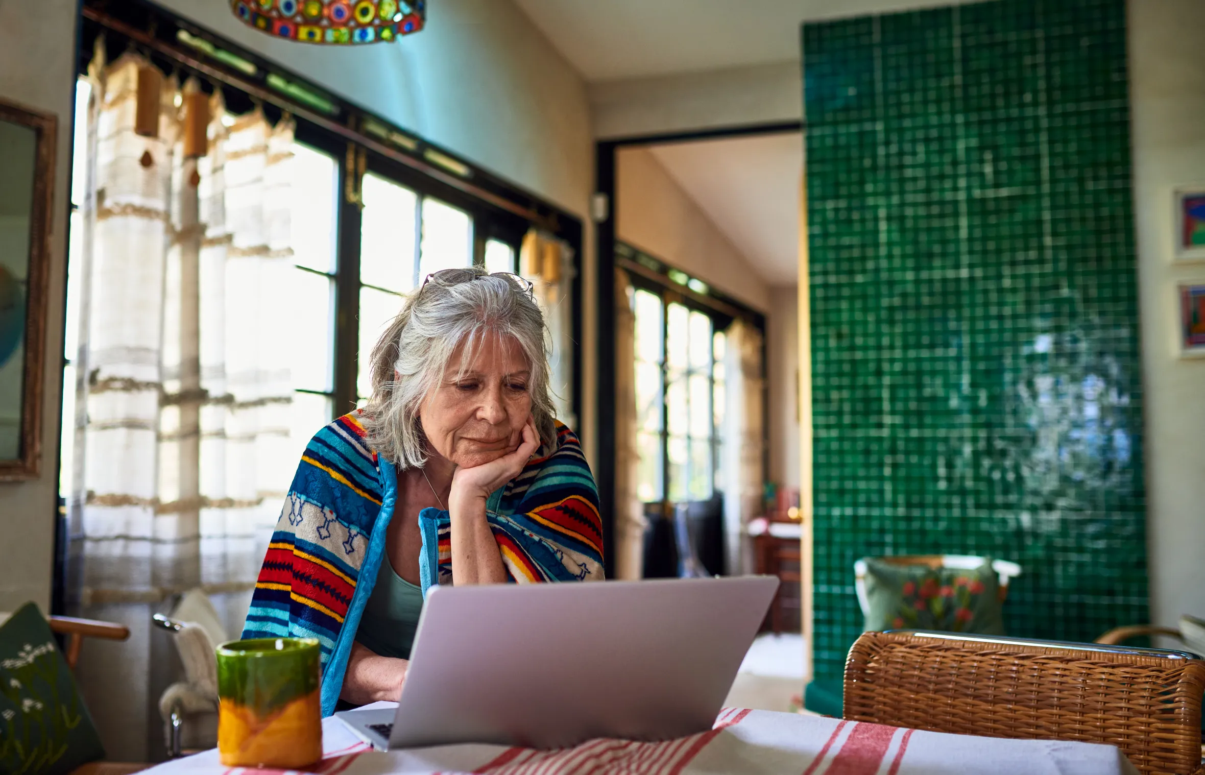Senior woman wrapped in a blanket, looking at a laptop at a dining table while working from home.